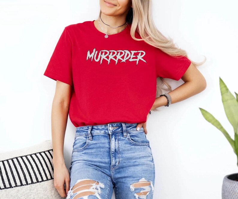 May include: A red t-shirt with the word "MURRRDER" in a spiky, silver font. The shirt is being worn with blue jeans. The person is standing in front of a white wall.