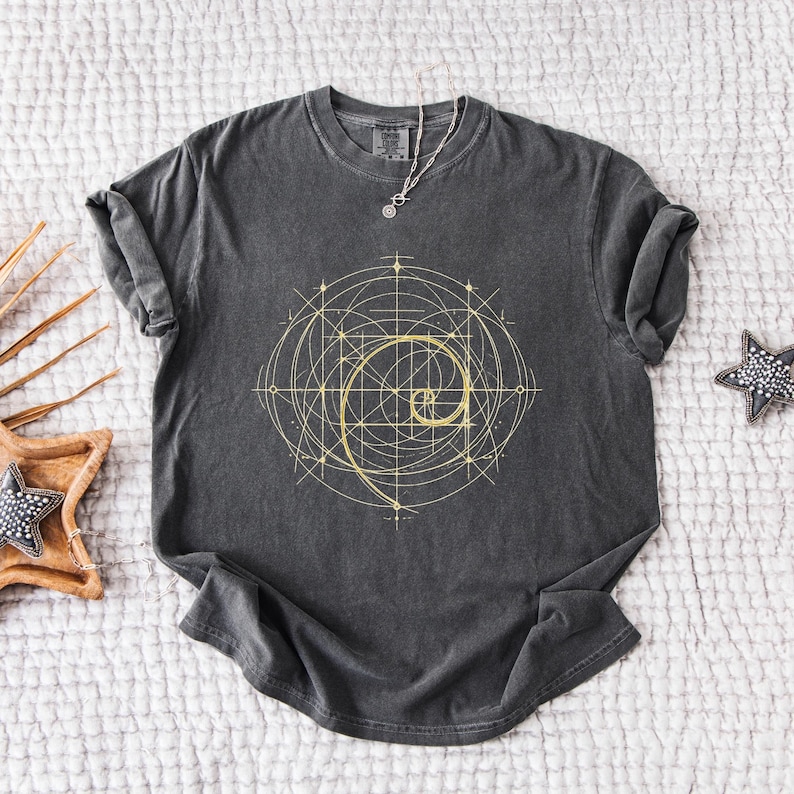 May include: Dark gray t-shirt featuring a golden spiral design with geometric lines. The shirt has a crew neck and short sleeves. A silver necklace with a pendant is draped on the shirt. The shirt is laying on a white surface.