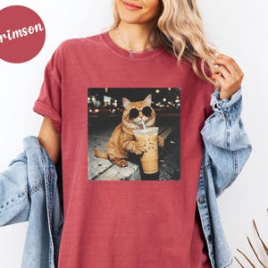 May include: Crimson-colored t-shirt featuring a square graphic of a cat wearing sunglasses and holding an iced coffee. The cat is orange and white, and the background is a blurred city scene. The shirt has short sleeves and a relaxed fit.