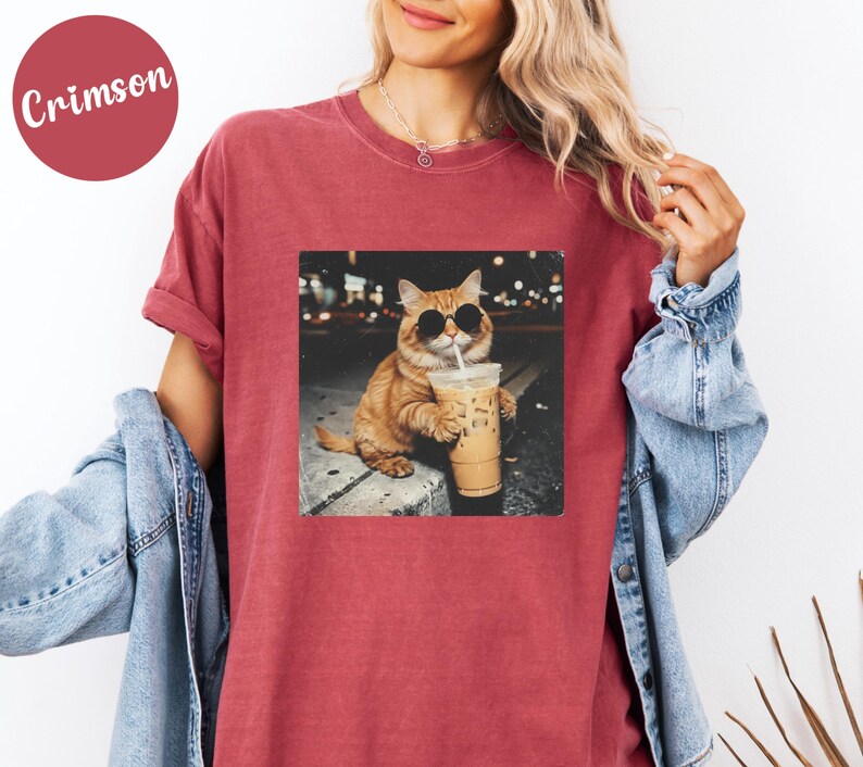 May include: Crimson-colored t-shirt featuring a square graphic of a cat wearing sunglasses and holding an iced coffee. The cat is orange and white, and the background is a blurred city scene. The shirt has short sleeves and a relaxed fit.