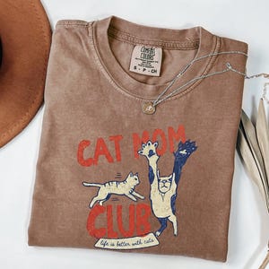 May include: A brown t-shirt with the words "CAT MOM CLUB" in red, and a graphic of two cats. The shirt also has the phrase "life is better with cats". A silver necklace with a pendant is on the shirt.