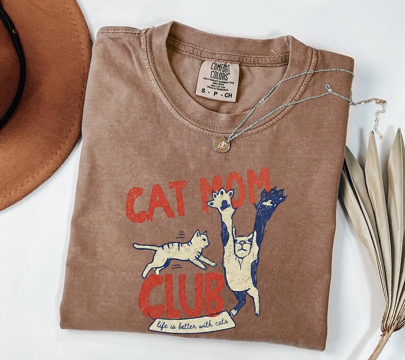 May include: A brown t-shirt with the words "CAT MOM CLUB" in red, and a graphic of two cats. The shirt also has the phrase "life is better with cats". A silver necklace with a pendant is on the shirt.