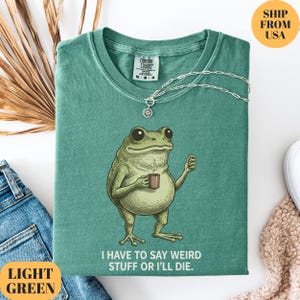 K&ouml;nnte beinhalten: Hellgr&uuml;nes T-Shirt mit einem Cartoon-Frosch, der eine Kaffeetasse h&auml;lt. Unter dem Frosch steht der Text "I HAVE TO SAY WEIRD STUFF OR I'LL DIE.". Eine silberne Halskette liegt &uuml;ber dem gefalteten T-Shirt.