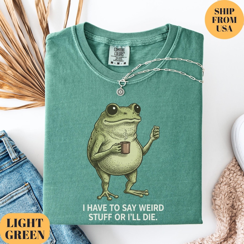 K&ouml;nnte beinhalten: Hellgr&uuml;nes T-Shirt mit einem Cartoon-Frosch, der eine Kaffeetasse h&auml;lt. Unter dem Frosch steht der Text "I HAVE TO SAY WEIRD STUFF OR I'LL DIE.". Eine silberne Halskette liegt &uuml;ber dem gefalteten T-Shirt.