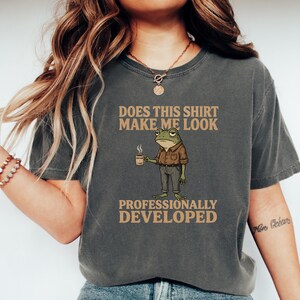 Peut inclure: Un t-shirt gris anthracite à manches courtes avec une illustration d'une grenouille verte portant une chemise marron et un pantalon gris, tenant une tasse de café. Le texte indique : DOES THIS SHIRT MAKE ME LOOK PROFESSIONALLY DEVELOPED.