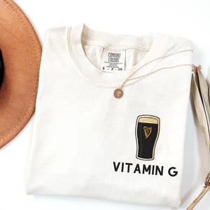 May include: Cream-colored t-shirt with a graphic of a pint of stout and the words "Vitamin G" in black. A gold necklace with a pendant rests on the shirt. The shirt is from Comfort Colors.