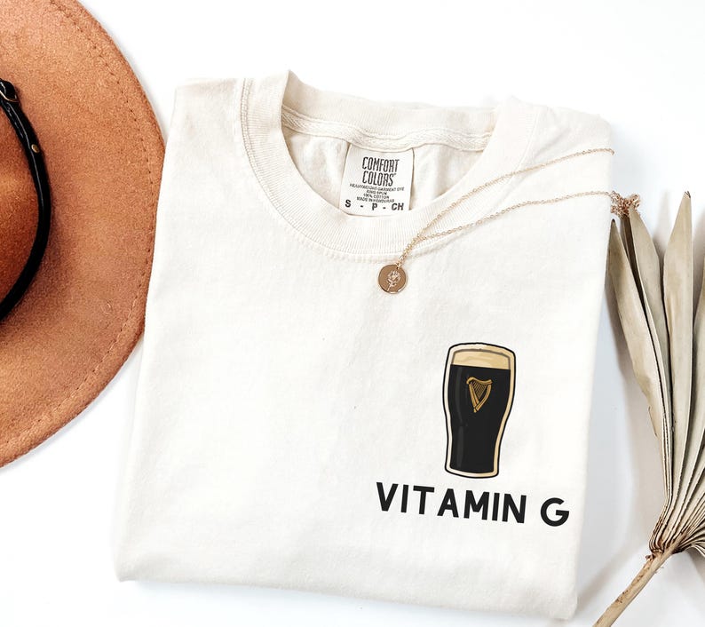 May include: Cream-colored t-shirt with a graphic of a pint of stout and the words "Vitamin G" in black. A gold necklace with a pendant rests on the shirt. The shirt is from Comfort Colors.