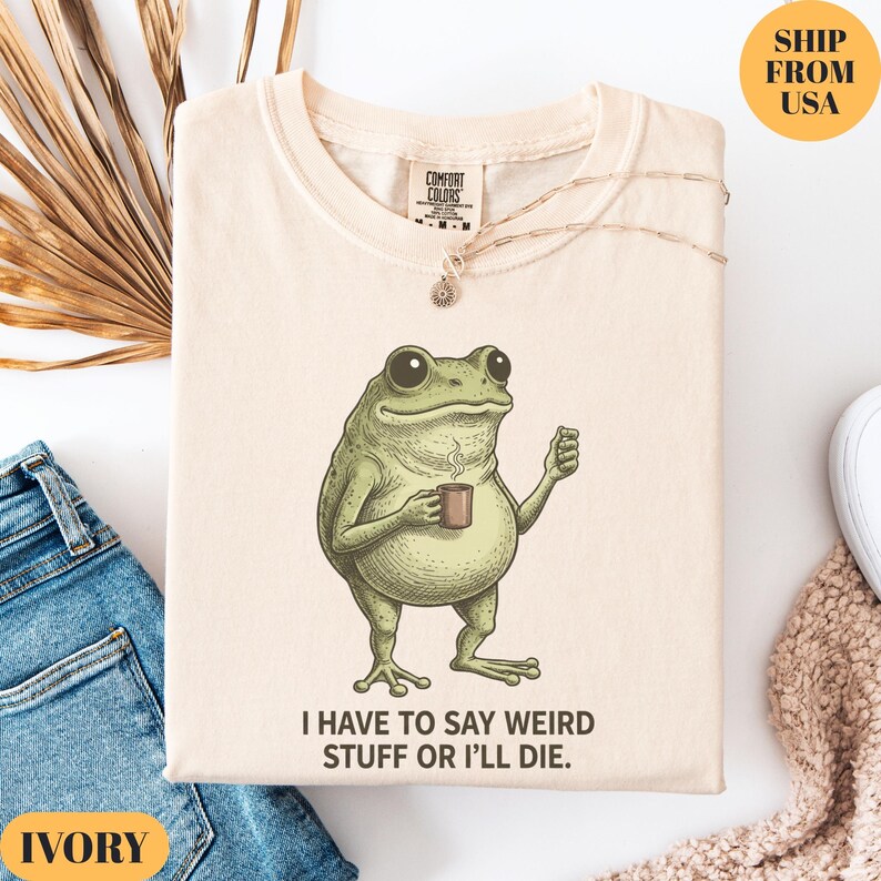 K&ouml;nnte beinhalten: Elfenbeinfarbenes T-Shirt mit einer Cartoon-Froschfigur, die eine Kaffeetasse h&auml;lt. Der Frosch ist gr&uuml;n mit gro&szlig;en Augen und steht aufrecht. Der Text "I HAVE TO SAY WEIRD STUFF OR I'LL DIE" ist darunter gedruckt. Eine goldene Halskette liegt auf dem Shirt.