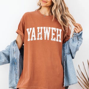 May include: A rust-colored t-shirt with the word "YAHWEH" printed in distressed white lettering. The shirt has short sleeves and a relaxed fit. A light blue denim jacket is draped over the shoulders.