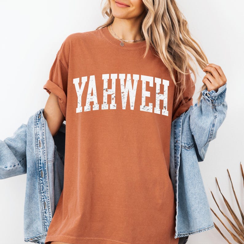 May include: A rust-colored t-shirt with the word "YAHWEH" printed in distressed white lettering. The shirt has short sleeves and a relaxed fit. A light blue denim jacket is draped over the shoulders.