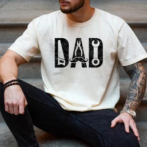 May include: A cream-coloured t-shirt featuring the word "DAD" in a large, distressed black font. The letters are cleverly designed with tools: a saw, pliers, a wrench, and a moustache. A casual, everyday wear item.