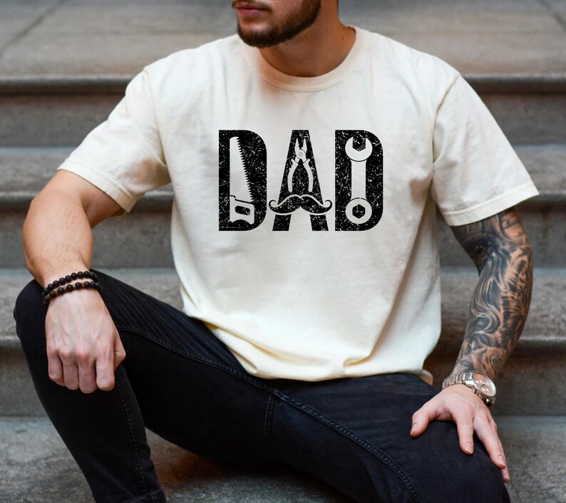 May include: A cream-coloured t-shirt featuring the word "DAD" in a large, distressed black font. The letters are cleverly designed with tools: a saw, pliers, a wrench, and a moustache. A casual, everyday wear item.