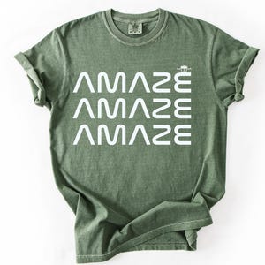 May include: An olive green t-shirt with the word "AMAZE" stacked three times in white, with a small graphic above the last word. The shirt has a crew neck and short sleeves.