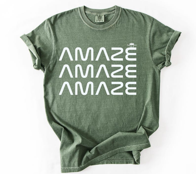 May include: An olive green t-shirt with the word "AMAZE" stacked three times in white, with a small graphic above the last word. The shirt has a crew neck and short sleeves.