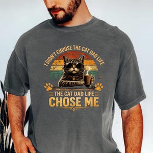 Op de afbeelding: Donkergrijs t-shirt met een afbeelding van een kat met een zonnebril en een koffiemok. De tekst luidt: "Ik koos niet voor het kattenvaderleven, het kattenvaderleven koos mij." Het ontwerp bevat pootafdrukken en een retro zonsondergang.