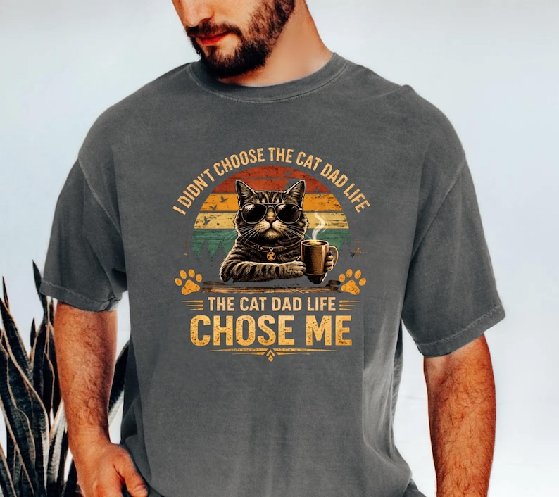 Op de afbeelding: Donkergrijs t-shirt met een afbeelding van een kat met een zonnebril en een koffiemok. De tekst luidt: "Ik koos niet voor het kattenvaderleven, het kattenvaderleven koos mij." Het ontwerp bevat pootafdrukken en een retro zonsondergang.