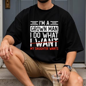 May include: Black t-shirt with the text "I'M A GROWN MAN I DO WHAT I WANT MY DAUGHTER WANTS" in white and red. The shirt is a casual style, suitable for everyday wear.