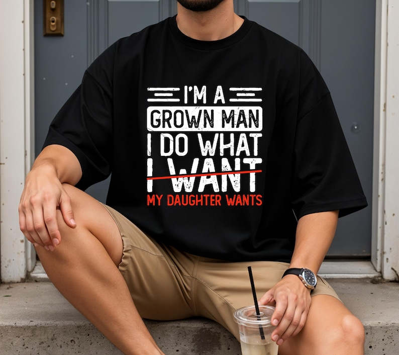 May include: Black t-shirt with the text "I'M A GROWN MAN I DO WHAT I WANT MY DAUGHTER WANTS" in white and red. The shirt is a casual style, suitable for everyday wear.