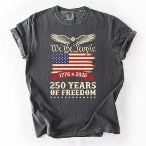May include: Dark grey t-shirt with a distressed American flag graphic and the text "We the People". Below the flag are the dates "1776 * 2026" and "250 YEARS OF FREEDOM". The design includes an eagle with outstretched wings.