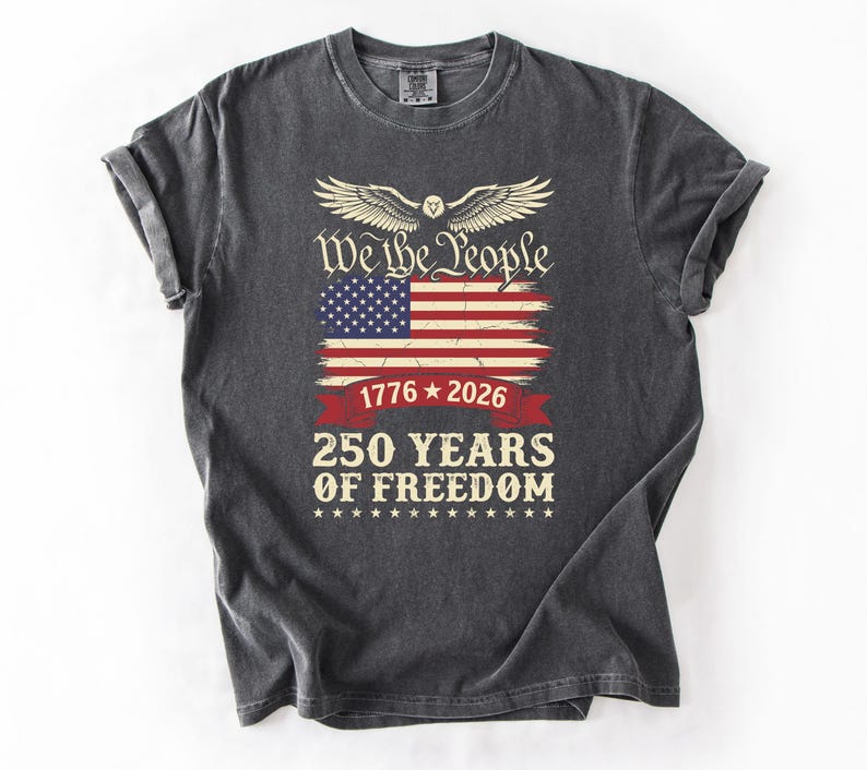 May include: Dark grey t-shirt with a distressed American flag graphic and the text "We the People". Below the flag are the dates "1776 * 2026" and "250 YEARS OF FREEDOM". The design includes an eagle with outstretched wings.