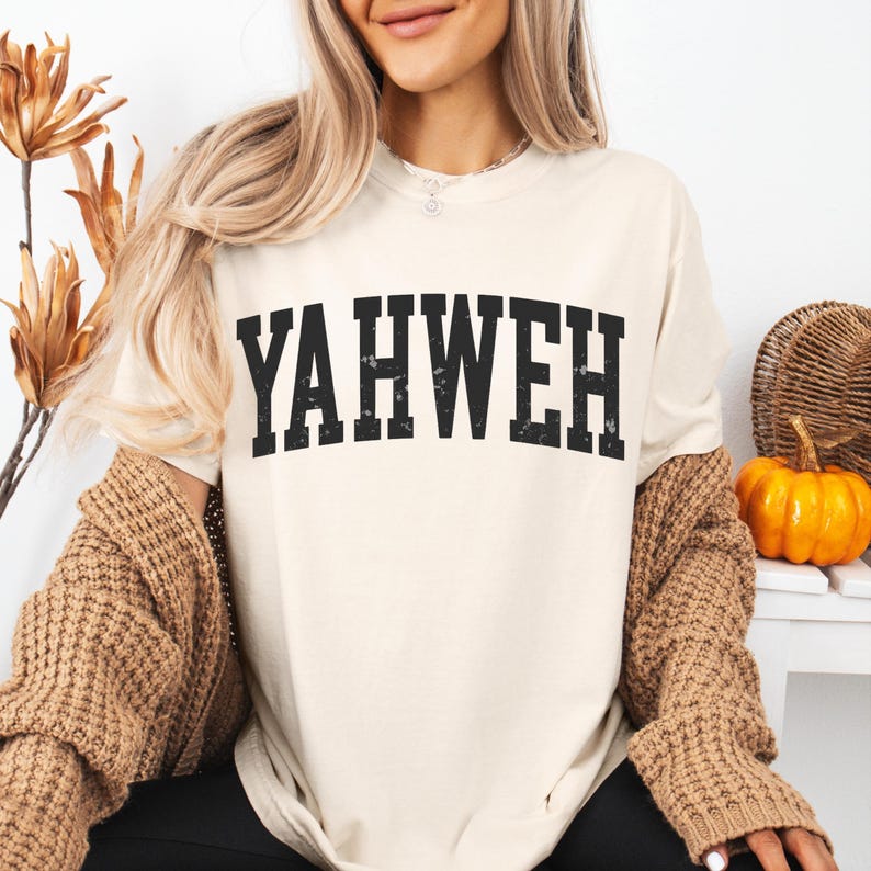 May include: Cream-colored t-shirt with the word "YAHWEH" printed in bold, black, distressed lettering. The shirt is paired with a brown cardigan. A small pumpkin and decorative elements are in the background.