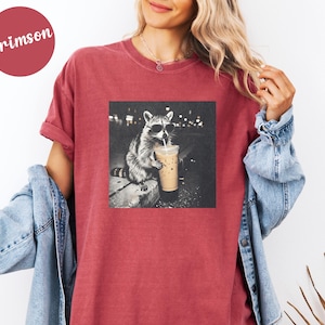 May include: Crimson-colored t-shirt featuring a black and white image of a raccoon wearing sunglasses and drinking from a coffee cup. The raccoon is sitting on a ledge. The shirt has a relaxed fit.