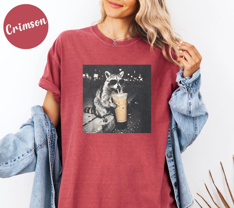 May include: Crimson-colored t-shirt featuring a black and white image of a raccoon wearing sunglasses and drinking from a coffee cup. The raccoon is sitting on a ledge. The shirt has a relaxed fit.