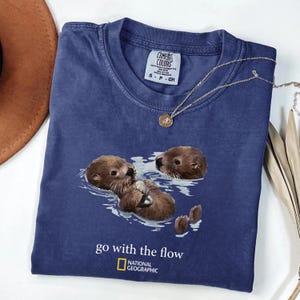 May include: A blue short-sleeved t-shirt featuring a graphic of two brown otters swimming in water. Below the otters, the text reads "go with the flow" and includes the National Geographic logo. The shirt has a Comfort Colors brand neck label.