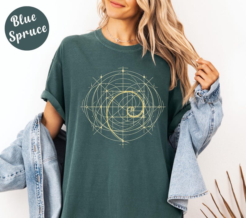 以下が含まれることがあります： ゴールデン幾何学模様のブルー・スプルースTシャツ。デザインには、スパイラルと交差する線が含まれています。「Blue Spruce」の文字が左上に円で表示されています。
