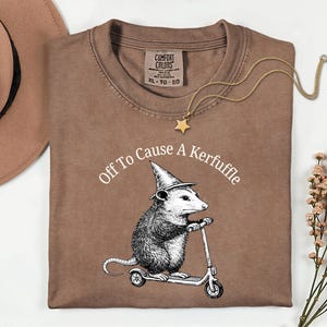 May include: A brown Comfort Colors t-shirt with the text "Off To Cause A Kerfuffle" above a graphic of an opossum wearing a witch hat and riding a scooter. A gold star pendant necklace is draped across the shirt.