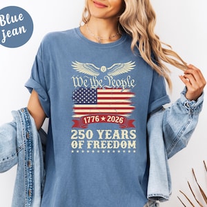 May include: Blue jean t-shirt featuring the American flag design with the text "We the People" and "250 Years of Freedom". The design includes the years 1776 and 2026. The shirt has short sleeves.