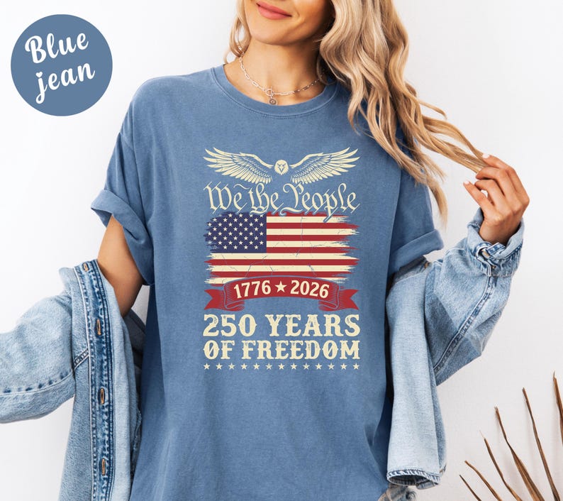 May include: Blue jean t-shirt featuring the American flag design with the text "We the People" and "250 Years of Freedom". The design includes the years 1776 and 2026. The shirt has short sleeves.