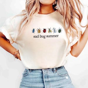 May include: Cream-colored t-shirt featuring a row of colorful bug illustrations above the text "sad bug summer" in black. The bugs are in blue, brown, red, light blue, and green. The shirt is made of a soft material.