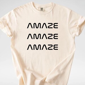 Pu&ograve; includere: T-shirt color crema con la parola "AMAZE" stampata tre volte in nero, impilata verticalmente. La maglietta ha maniche corte e girocollo. Il tessuto sembra morbido e confortevole.