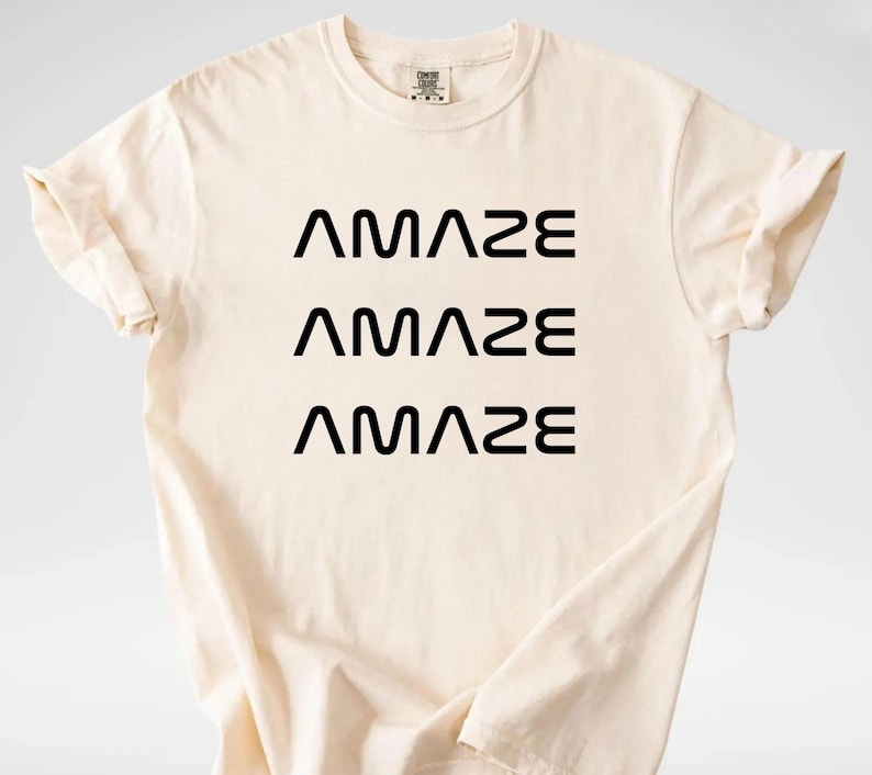 Pu&ograve; includere: T-shirt color crema con la parola "AMAZE" stampata tre volte in nero, impilata verticalmente. La maglietta ha maniche corte e girocollo. Il tessuto sembra morbido e confortevole.