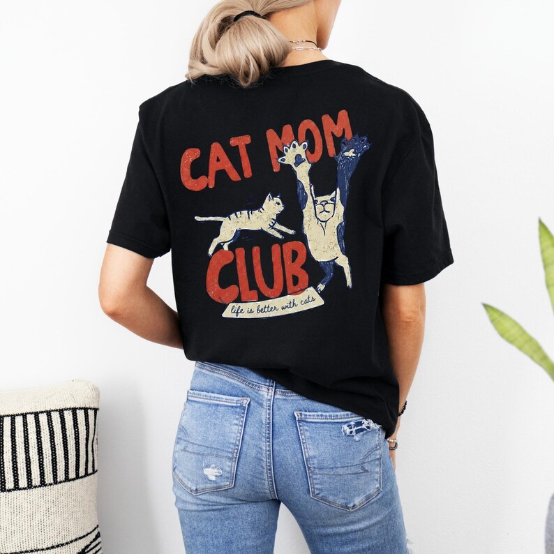 May include: Black t-shirt with a distressed graphic that reads "CAT MOM CLUB" in red and white. The design features two cats, one running and the other with its paws up. The text "life is better with cats" is below the word "CLUB".