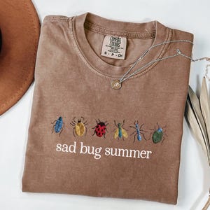 May include: A brown t-shirt with embroidered bugs in various colors and the text "sad bug summer". A silver necklace with a pendant is draped across the shirt. The shirt is folded on a white surface.