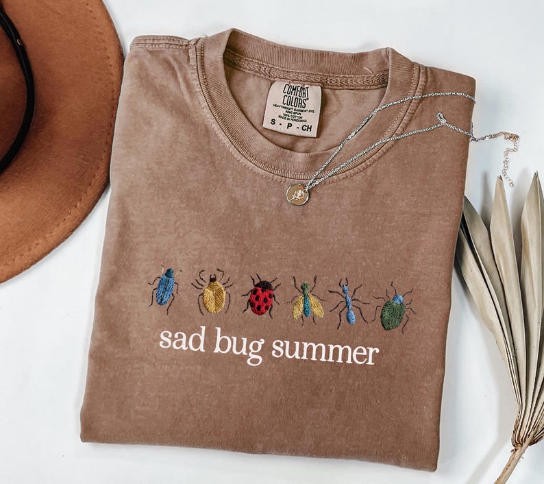 May include: A brown t-shirt with embroidered bugs in various colors and the text "sad bug summer". A silver necklace with a pendant is draped across the shirt. The shirt is folded on a white surface.