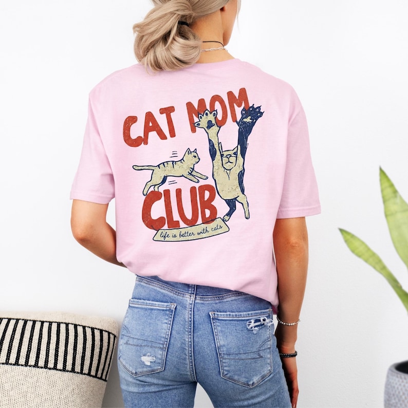May include: Light pink t-shirt with a graphic design featuring the words "CAT MOM CLUB" in red and a cartoon cat illustration. The phrase "life is better with cats" is printed below the club name.