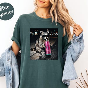 May include: Blue Spruce t-shirt featuring a graphic of a raccoon wearing sunglasses and drinking from a pink tumbler. The raccoon is sitting on a curb with a city background. The shirt is a solid teal color.