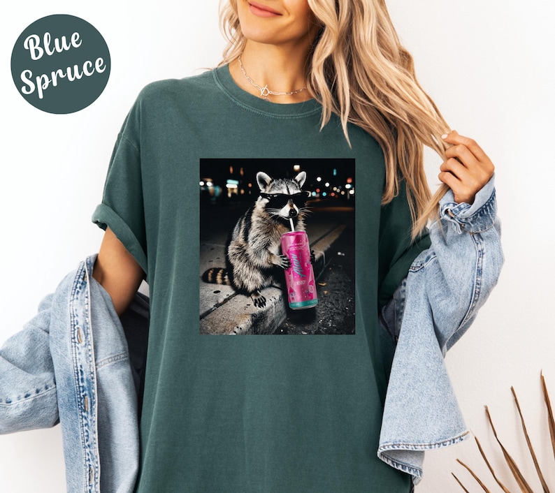 May include: Blue Spruce t-shirt featuring a graphic of a raccoon wearing sunglasses and drinking from a pink tumbler. The raccoon is sitting on a curb with a city background. The shirt is a solid teal color.
