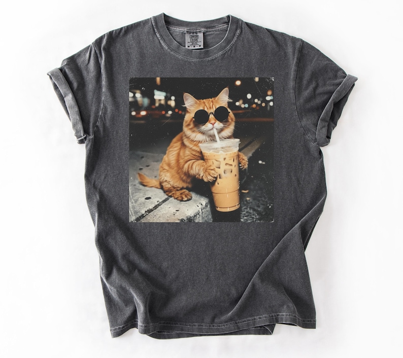 May include: Dark gray t-shirt featuring a graphic of an orange cat wearing sunglasses and drinking an iced coffee. The cat is sitting on a ledge with a blurred city background. The shirt is made of a soft material.