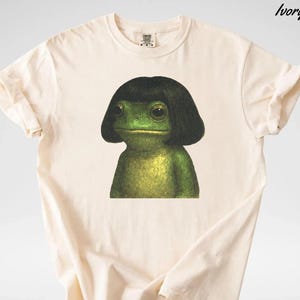 May include: Ivory-colored t-shirt featuring a green frog with a black wig. The frog has large eyes and a neutral expression. The shirt is made of a soft material and has a classic crew neck design.