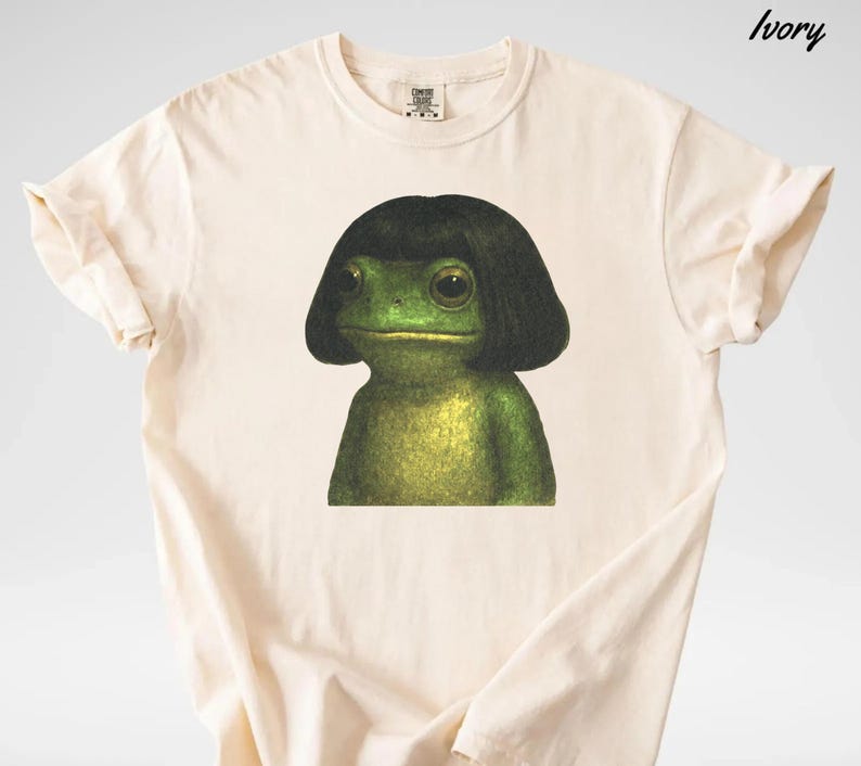 May include: Ivory-colored t-shirt featuring a green frog with a black wig. The frog has large eyes and a neutral expression. The shirt is made of a soft material and has a classic crew neck design.