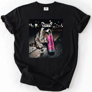 May include: Black t-shirt featuring a raccoon wearing sunglasses and drinking from a pink can with a straw. The raccoon is sitting on a concrete ledge with a blurred city background. The shirt has a classic crew neck.