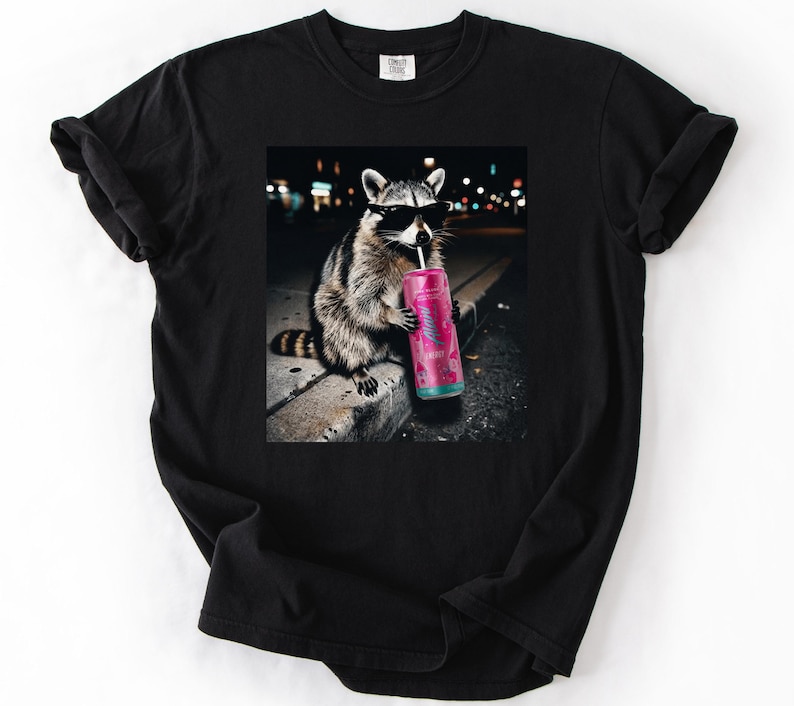 May include: Black t-shirt featuring a raccoon wearing sunglasses and drinking from a pink can with a straw. The raccoon is sitting on a concrete ledge with a blurred city background. The shirt has a classic crew neck.