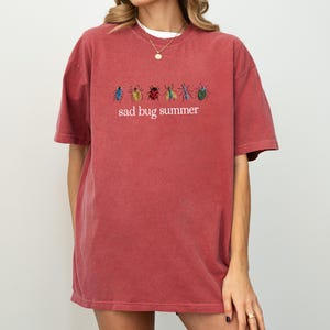 May include: A red-orange t-shirt with the embroidered text "sad bug summer" and colorful bug illustrations. The shirt has a crew neck and short sleeves. The fabric appears soft and slightly faded.