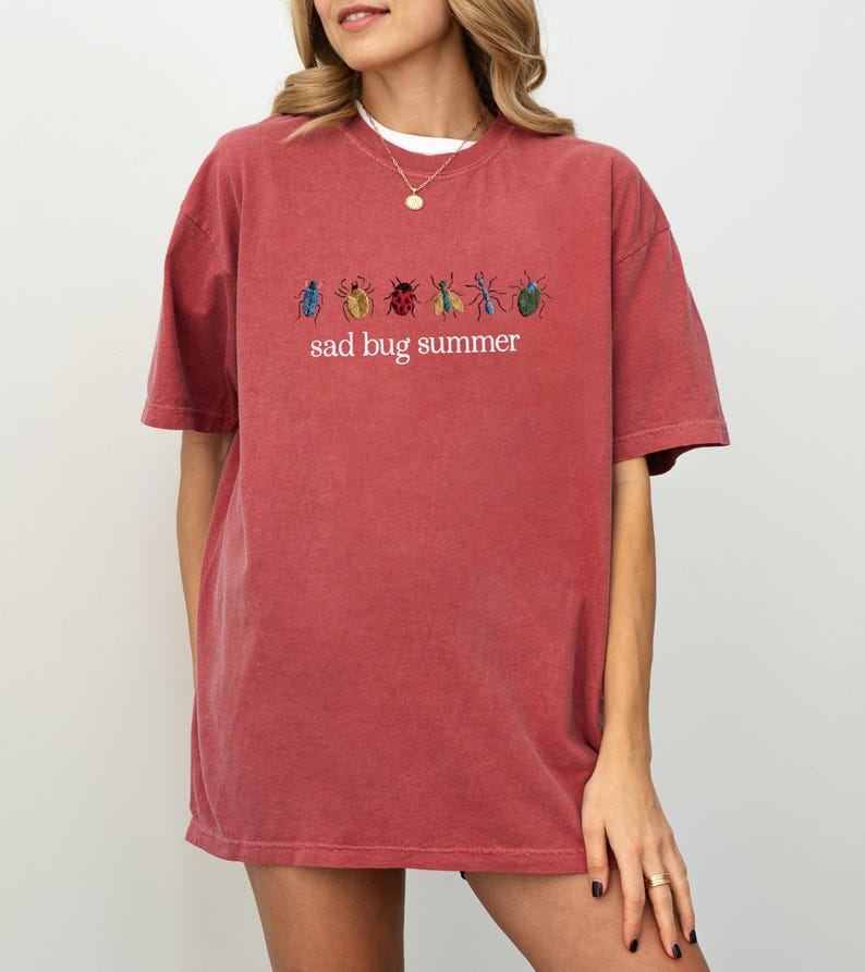 May include: A red-orange t-shirt with the embroidered text "sad bug summer" and colorful bug illustrations. The shirt has a crew neck and short sleeves. The fabric appears soft and slightly faded.