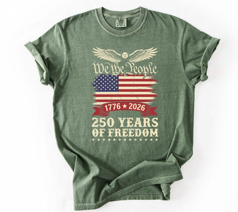 May include: A green t-shirt with a graphic design featuring the American flag, the text "We the People," and "250 Years of Freedom." The design includes the years 1776 and 2026, and a banner.
