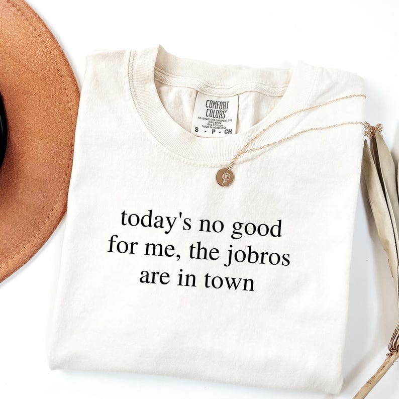 Op de afbeelding: Cr&egrave;mekleurig T-shirt met de zwarte tekst "today's no good for me, the jobros are in town". Een gouden ketting met hanger en een bruine hoed liggen naast het shirt.